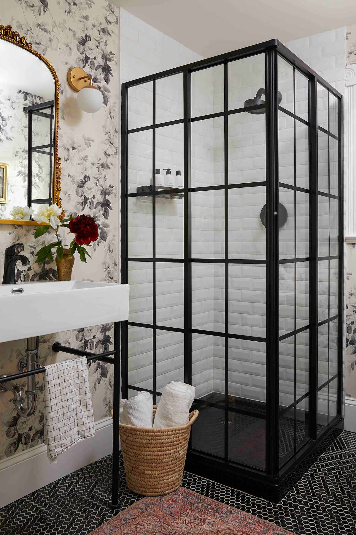 A chic bathroom features a glass-enclosed shower with black framing, showcasing white subway tiles. A vintage-style mirror hangs above the sink, with floral wallpaper providing a soft backdrop. A woven basket holds neatly rolled towels on the floor, adding warmth to the space.