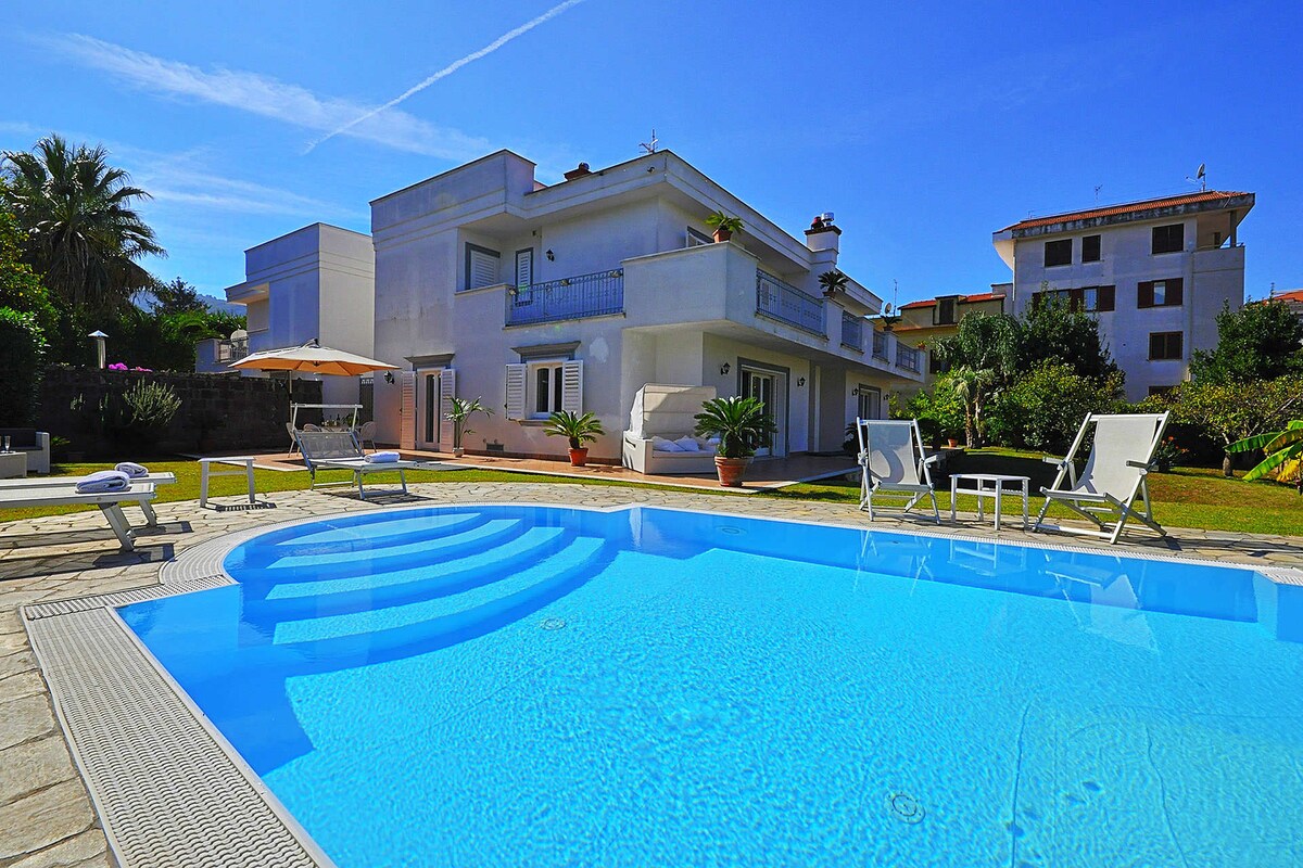 The image captures a sunny outdoor space featuring a private swimming pool. Sun loungers are positioned beside the pool, offering relaxation. The villa is visible in the background, showcasing a modern architectural design. Lush greenery surrounds the property, contributing to the tranquil setting.