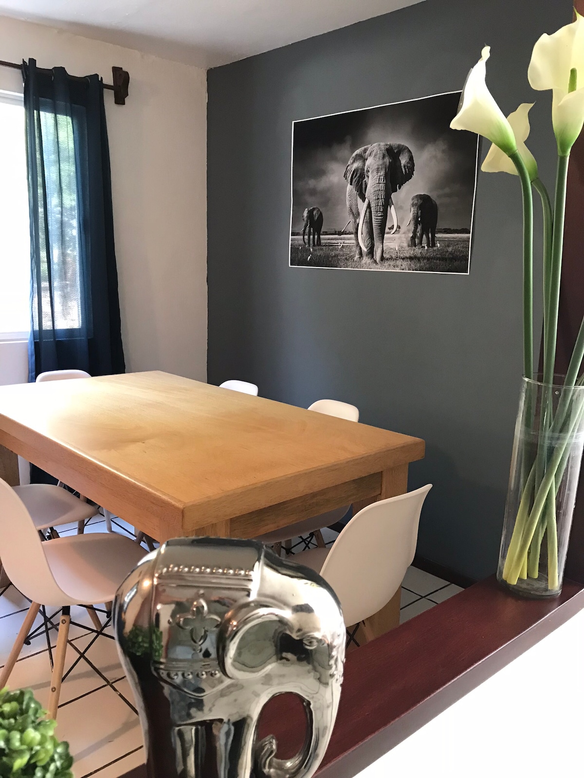 A dining area features a wooden table surrounded by six white chairs, illuminated by natural light from a nearby window. A large black-and-white photograph of elephants adorns the wall, complemented by tall white flowers in a glass vase and decorative greenery on the table.