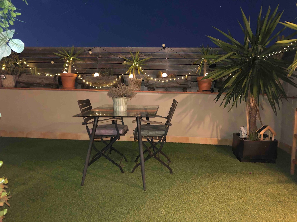 A private terrace is viewed at night, featuring a glass table with two folding chairs. Potted plants and soft lighting create a calming atmosphere, accentuating the greenery of the artificial lawn in the space.