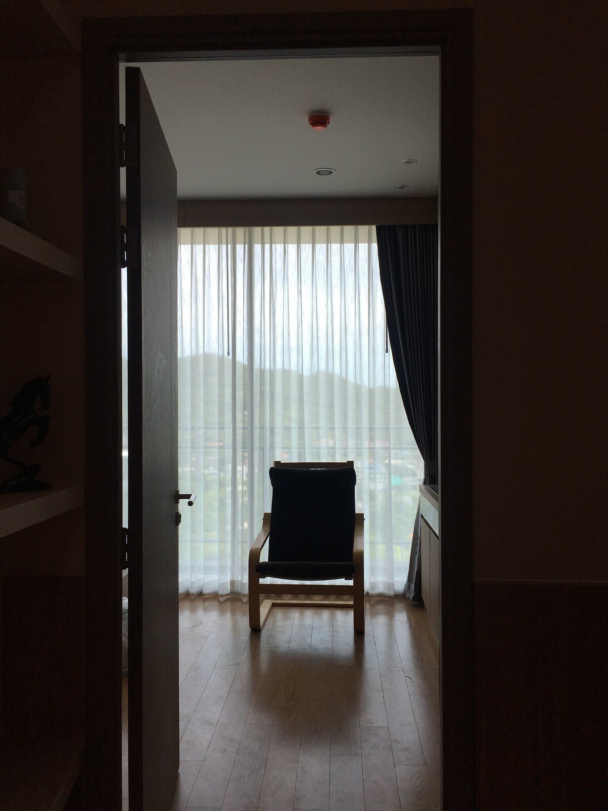 A doorway leads into a room with a wooden floor. A tall window with sheer curtains allows soft light to enter, revealing a view of distant hills. A single wooden chair is positioned facing the window, inviting relaxation.