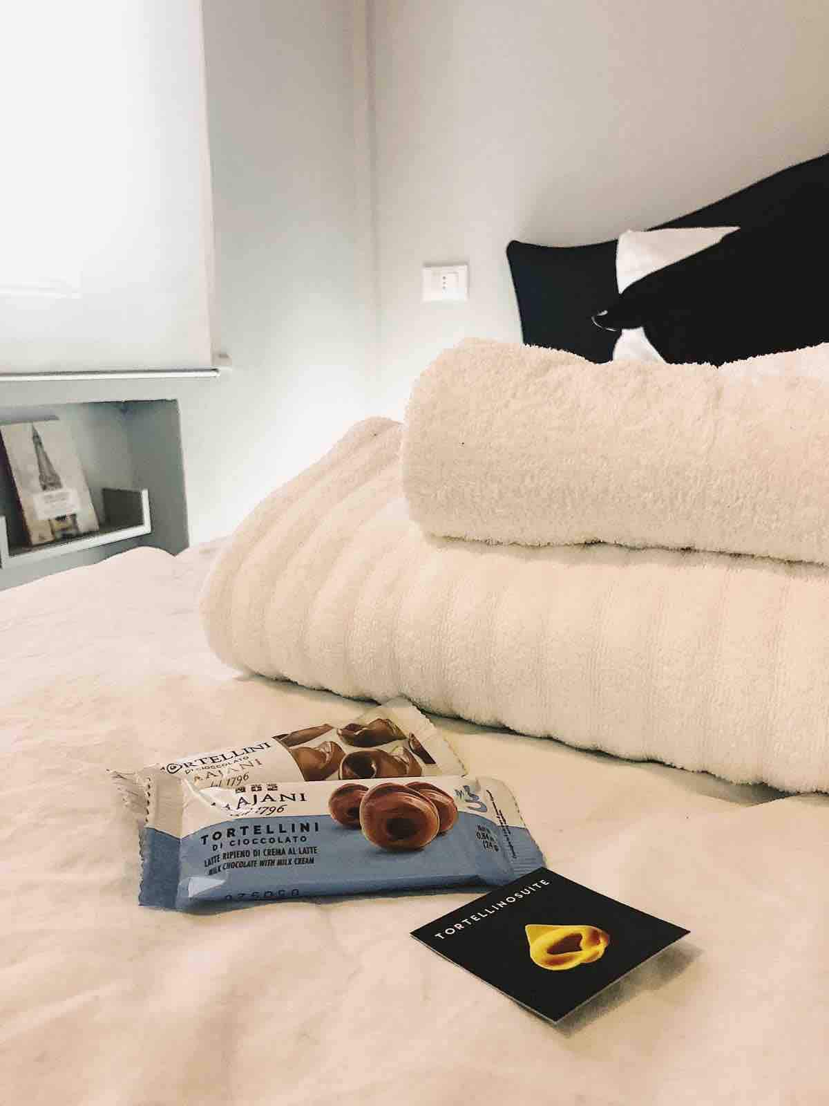 A neatly arranged bed is complemented by neatly stacked white towels. A small pack of tortellini and a brochure are displayed on the bed, inviting guests to explore local culinary delights.