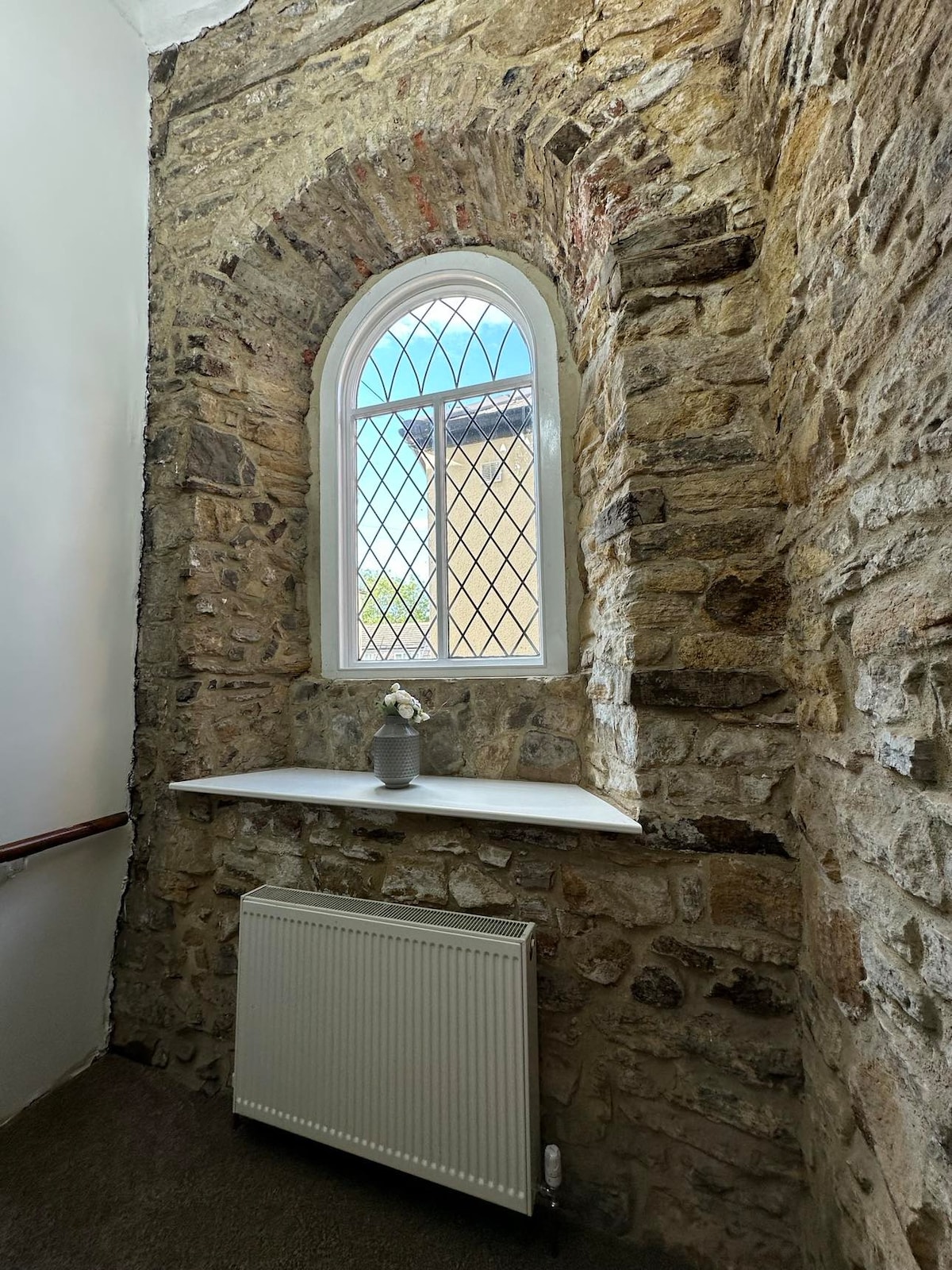 A stone wall staircase area features an arched window with decorative glass, allowing natural light to enter. A small ledge with a decorative plant sits beneath the window, while a radiator is positioned against the wall. The original stone highlights the historical character of the space.