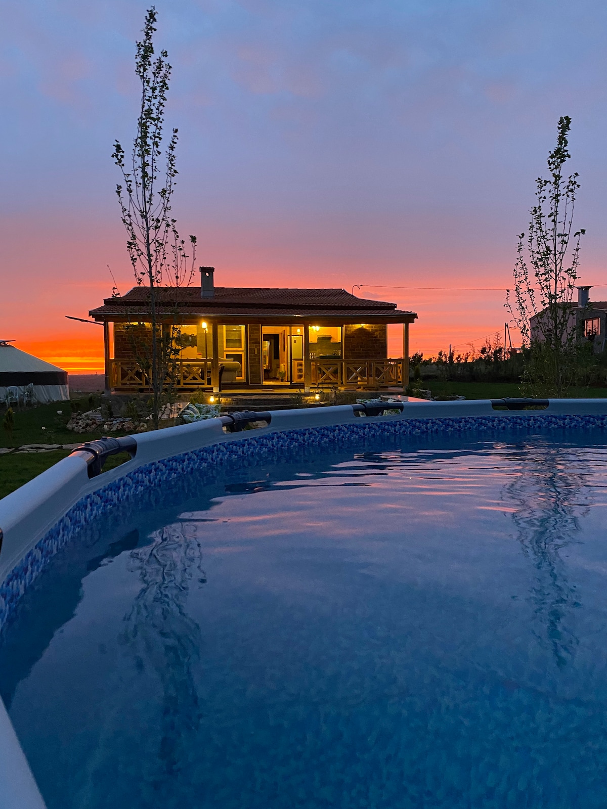 A calm scene features a shimmering pool reflecting the hues of a sunset sky. The chalet is showcased in the background, illuminated warmly, with a welcoming ambiance created by the lighting. Tall trees frame the edges of the photo, enhancing the peaceful setting.