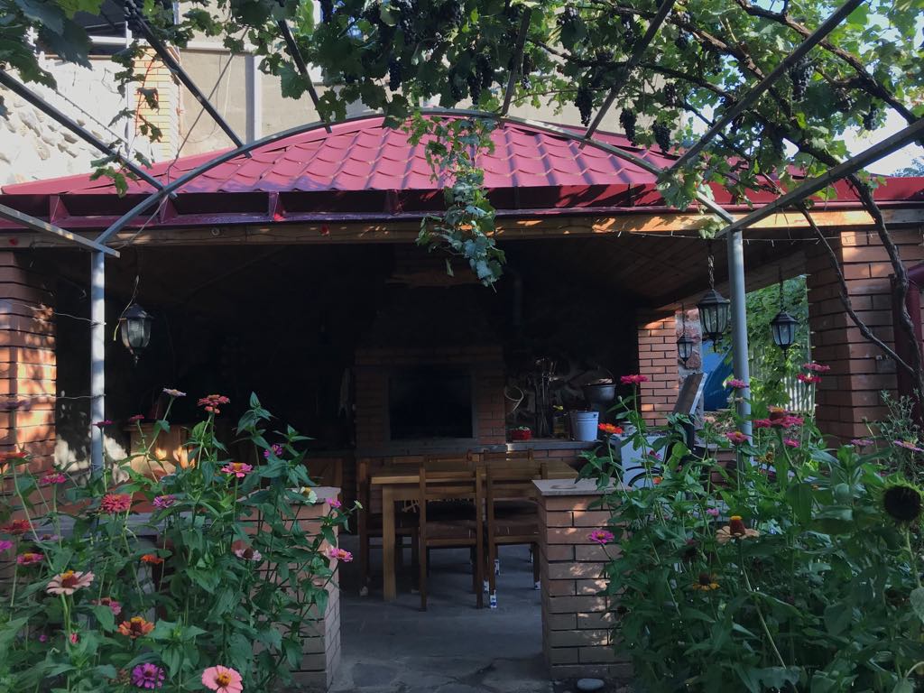 A covered outdoor seating area is surrounded by blooming flowers and greenery. The structure features wooden tables and chairs. Lanterns hang from the roof, offering a cozy ambiance. A stone barbecue grill is visible, providing a space for outdoor cooking.