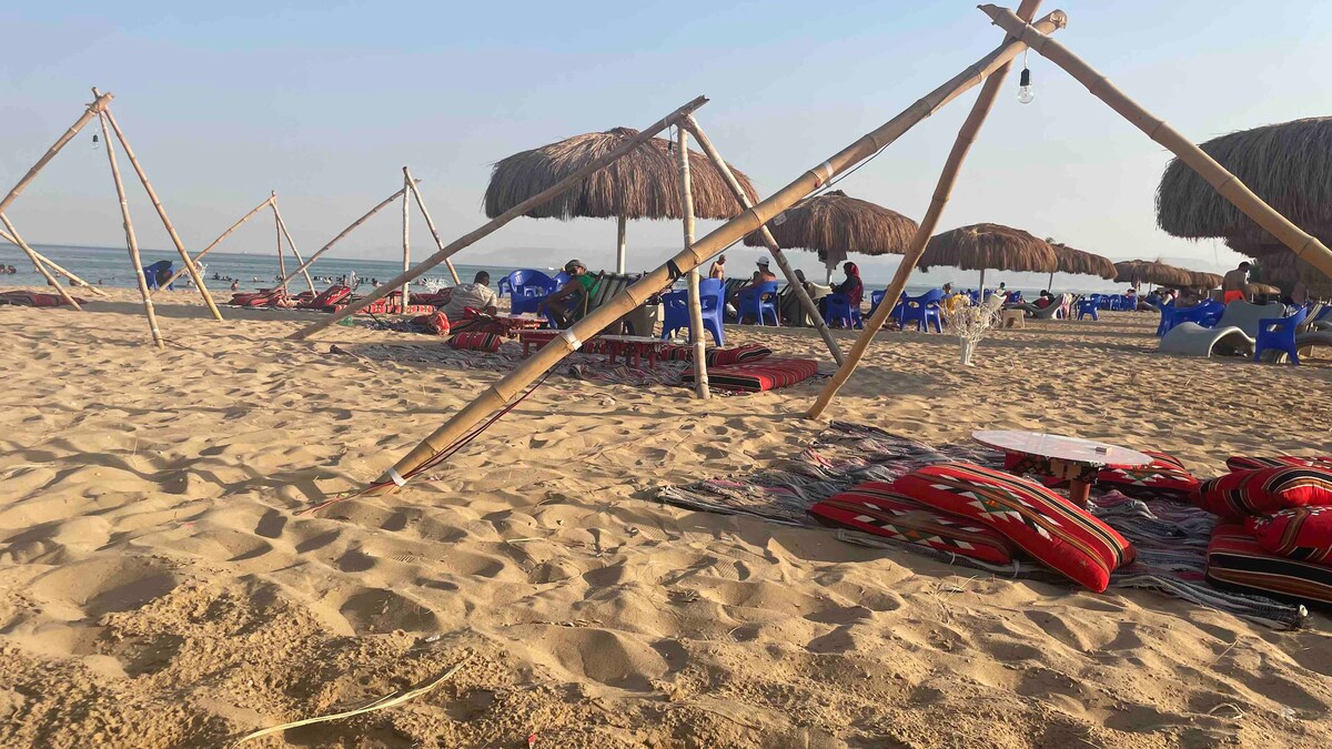 A sandy beach scene features several straw umbrellas providing shade. Colorful lounge mats and chairs are arranged in the sand, creating a relaxed atmosphere. The gentle waves of the sea are visible in the background, enhancing the beach setting.