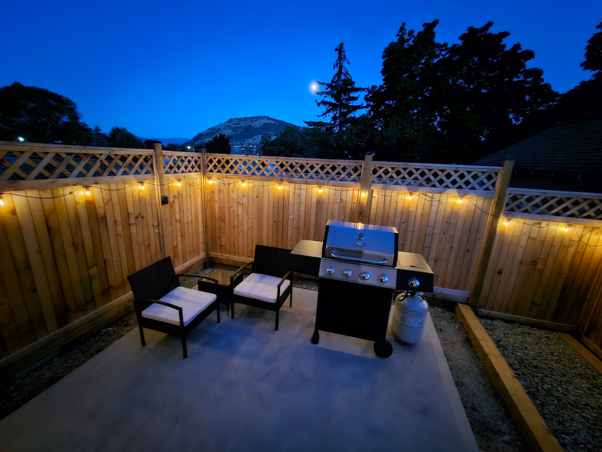 An outdoor patio area is presented with two comfortable chairs and a grill, creating a space for relaxation and cooking. Soft lighting adorns the wooden fence, while trees and a mountain silhouette are visible in the background against the evening sky.