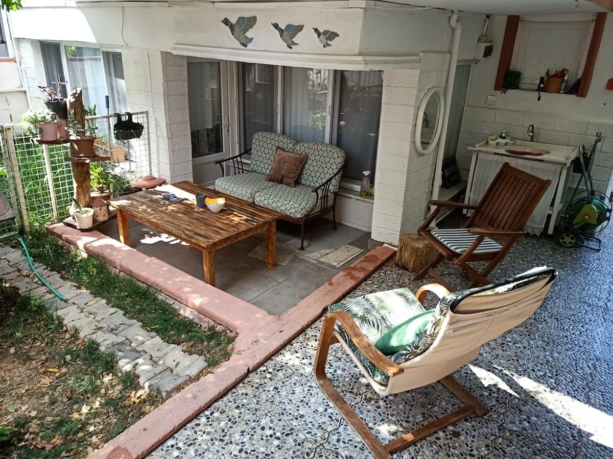 A shaded outdoor seating area features a wooden coffee table positioned between two comfortable chairs. A patterned seating arrangement complements the rustic ambiance. Potted plants add greenery to the space, enhancing the inviting atmosphere for relaxation.