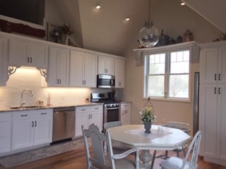 A modern kitchen features white cabinetry and stainless steel appliances. The space is illuminated by natural light from a large window. A simple round table is set for dining, adorned with a small floral arrangement, complementing the inviting atmosphere.