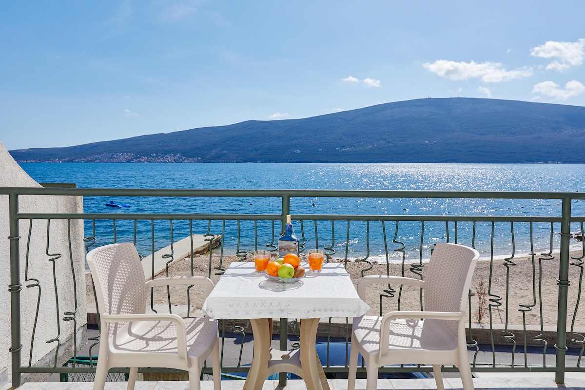A tranquil balcony is featured, overlooking a serene sea. A small table is set with fruit and drinks, accompanied by two white chairs. Beyond, the calm water sparkles under clear blue skies, with a distant mountain visible on the horizon.