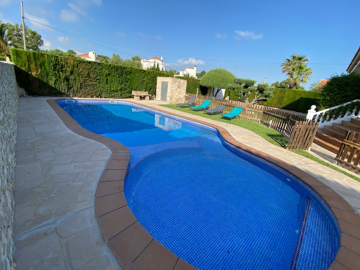 A spacious outdoor area features a large swimming pool surrounded by sun loungers. Lush greenery lines the property, providing a sense of privacy. The pool's clear blue water is complemented by the sunlit stone patio, creating an inviting space for relaxation.