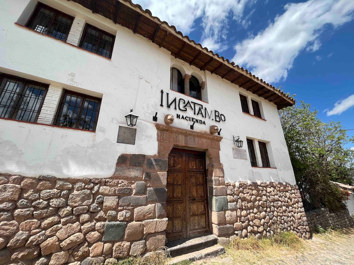 The exterior of the hacienda features a sturdy wooden door framed by a stone façade. Text indicating 'Hacienda Inka Tambo' is displayed prominently above the entrance. Large windows with wrought iron bars are visible, surrounded by natural stone and wooden beam accents.