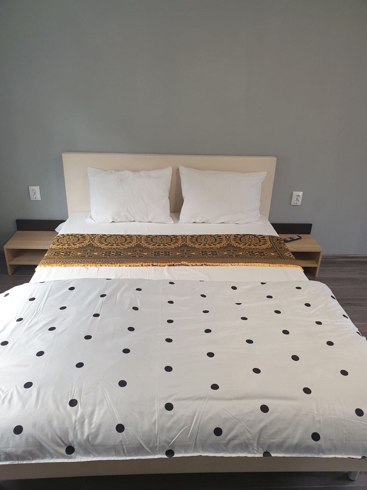 A spacious bed is presented with white pillows and a patterned throw blanket. The bedding features a design with black polka dots, complementing the neutral-colored walls. Nightstands are placed on either side of the bed, equipped with power outlets for convenience.