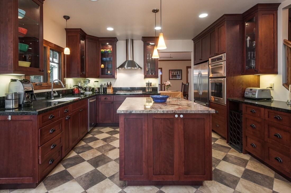 A spacious kitchen is designed with rich wood cabinetry and a large central island featuring a granite countertop. Modern appliances, including a stainless-steel oven and hood, are complemented by decorative overhead lighting. The tile floor showcases a checkerboard pattern, adding visual interest to the room.