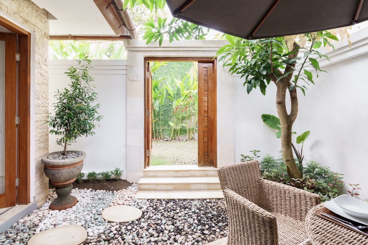 An outdoor area features a small seating arrangement with woven chairs and a table under a large umbrella. Smooth stones and pebbles create a path leading to a wooden door framed by lush greenery and tropical plants, adding a sense of natural tranquility.