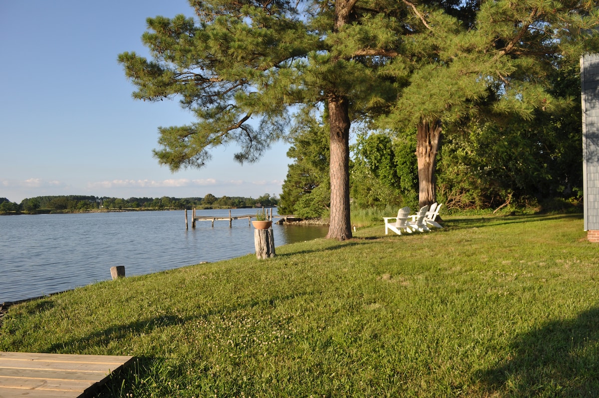 Lush green grass leads to the waterfront, where the calm water reflects the surroundings. Two white Adirondack chairs sit invitingly under the shade of tall trees, providing a serene spot for relaxation. A wooden dock extends into the water, enhancing the picturesque setting.
