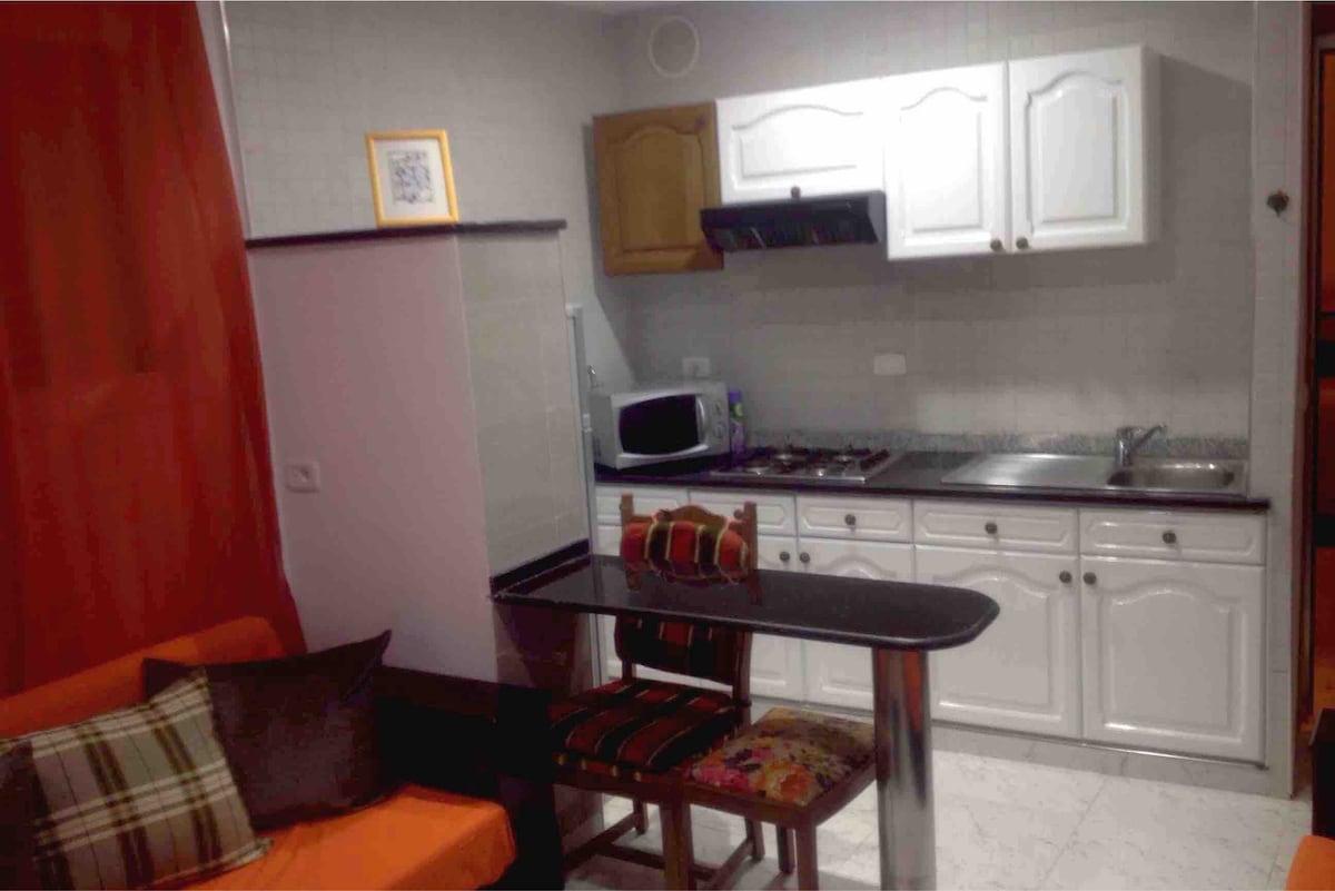 A compact kitchenette features white cabinetry and appliances, including a microwave and a stovetop. A small dining table with colorful seating is positioned nearby. An orange sofa adds a pop of color to the space, while a red curtain provides privacy.