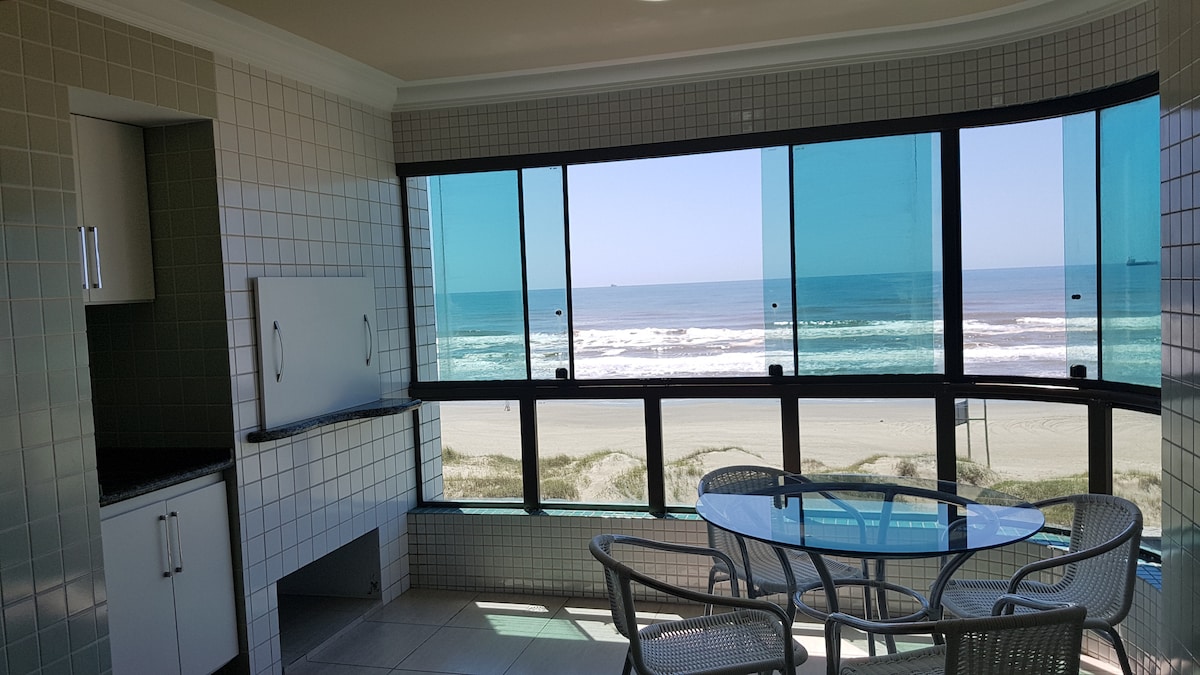 A bright dining area features a glass table and four chairs, set against large windows offering an unobstructed view of the ocean. Natural light floods the room, highlighting the tiled walls and the kitchenette adjacent to the dining space.
