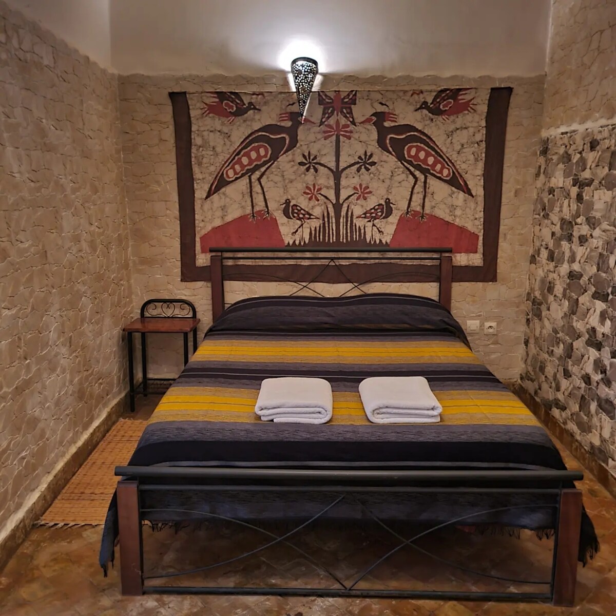 A cozy bedroom is featured, showcasing a spacious bed adorned with a dark striped blanket. Two neatly folded towels are placed at the foot of the bed. The wall behind is decorated with a colorful textile art piece, surrounded by stone-textured walls, contributing to a serene ambiance.