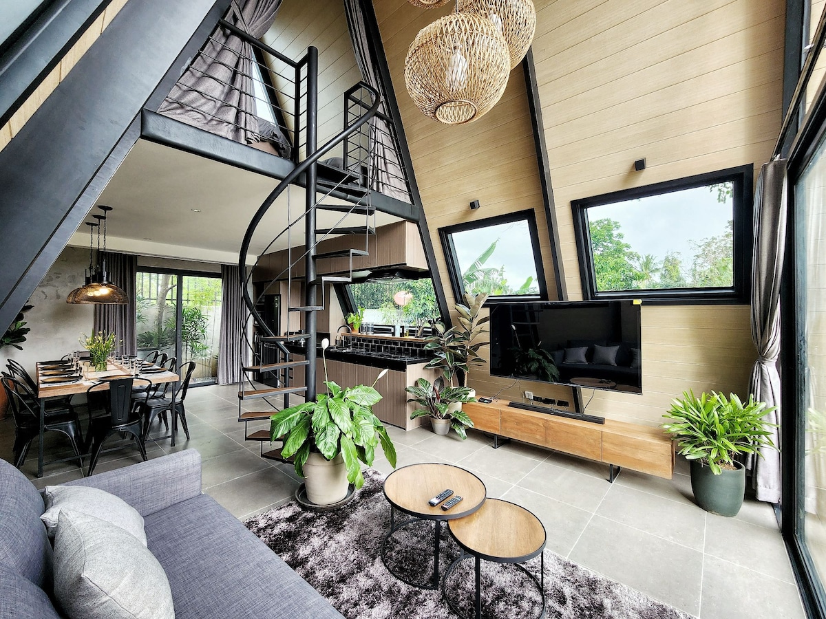 An open living area features a spiral staircase leading to the upper level. Large windows allow natural light to illuminate the space, while tropical plants adorn the interior. A comfortable sofa faces a media console with a television, complemented by a dining area in the background.