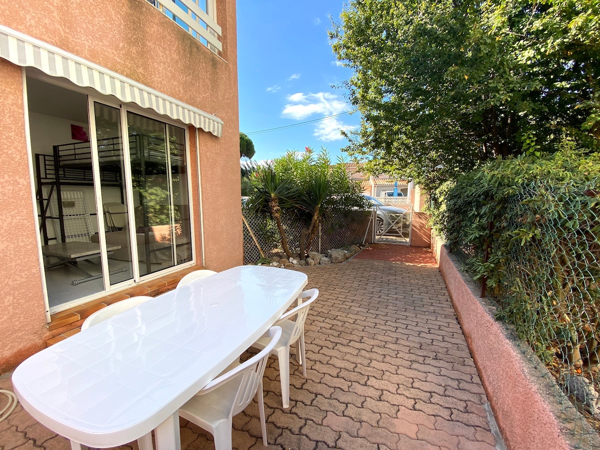 A spacious terrace area is shown, featuring a large white table surrounded by several white chairs. The space is partially shaded, bordered by greenery and fencing, with sunlight filtering through trees and a clear blue sky visible above.