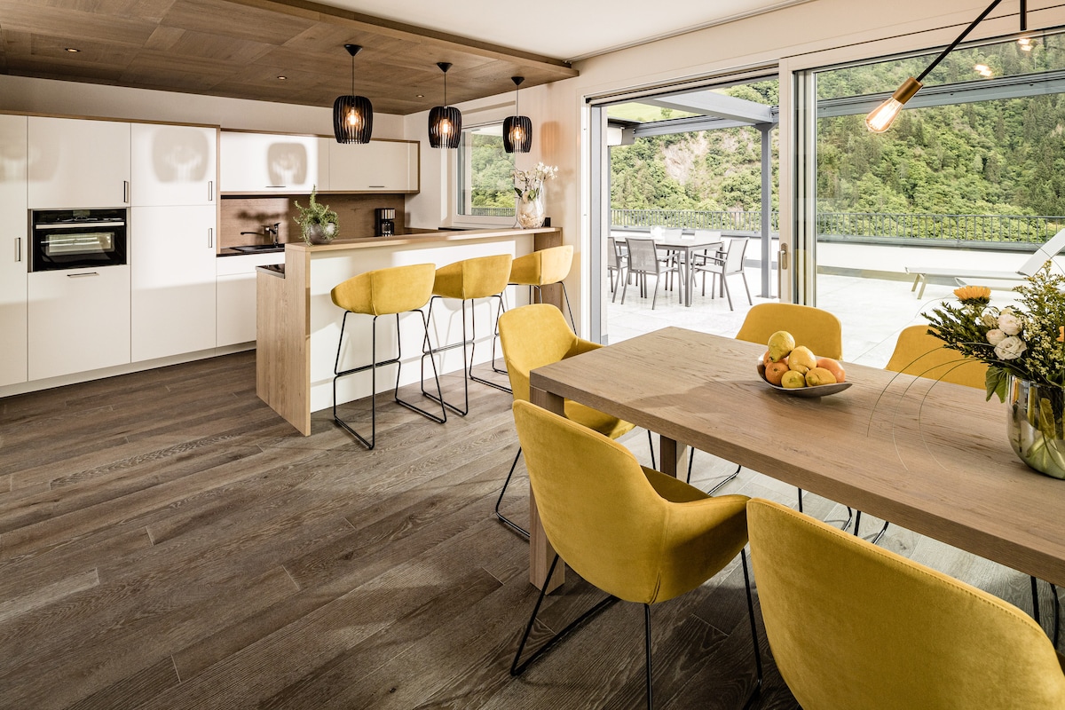 The open-concept living area displays a modern kitchen with stylish cabinetry and a spacious dining table surrounded by bright yellow chairs. Large glass doors provide a view of the outdoor terrace, enhancing the natural light within the space.