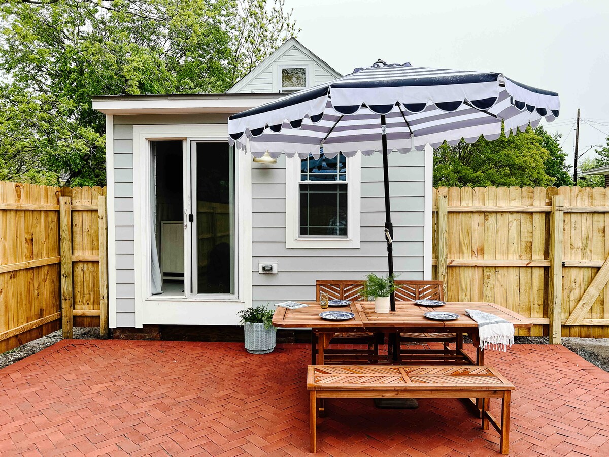 A fenced patio area features a wooden dining table with benches, set for outdoor meals. A large striped umbrella provides shade. The house in the background shows sliding glass doors leading into the interior, complemented by a nearby potted plant.