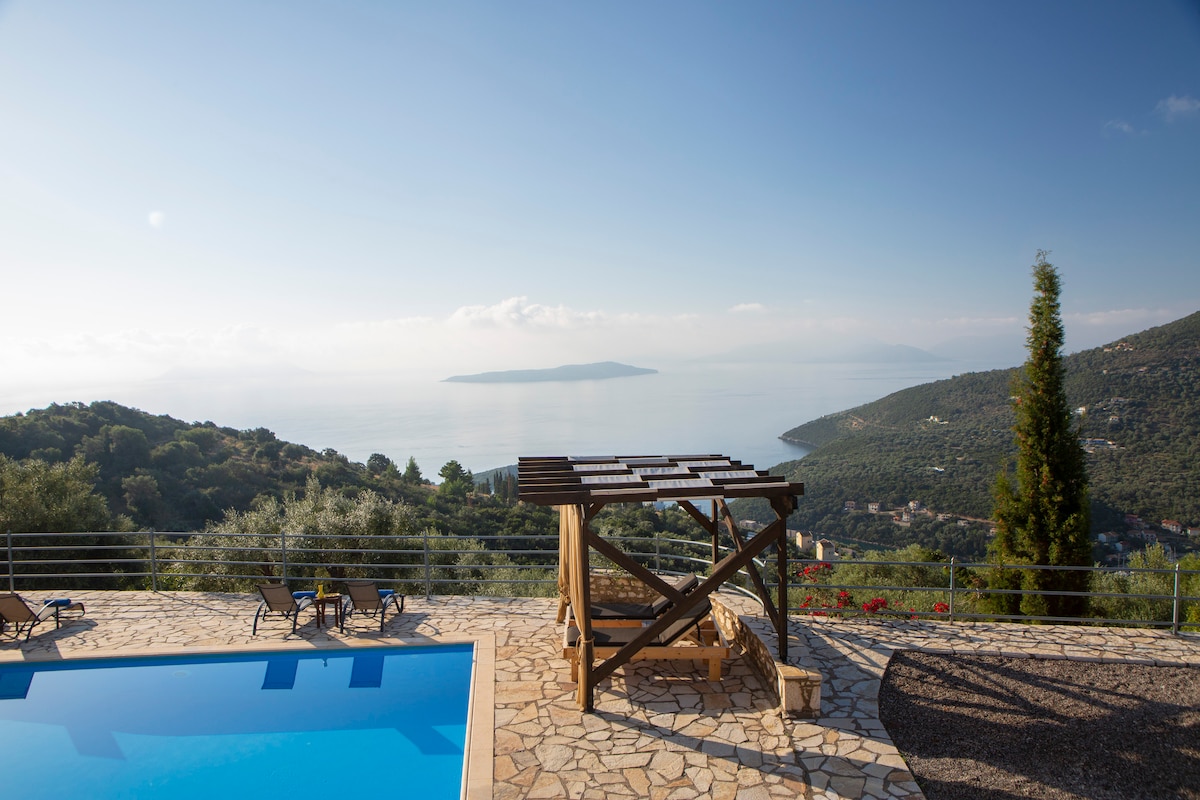 A tranquil outdoor space features a blue pool with sun loungers arranged around it. A shaded pergola stands nearby, offering views of the distant hills and sea. Lush greenery surrounds the area, complementing the serene landscape.
