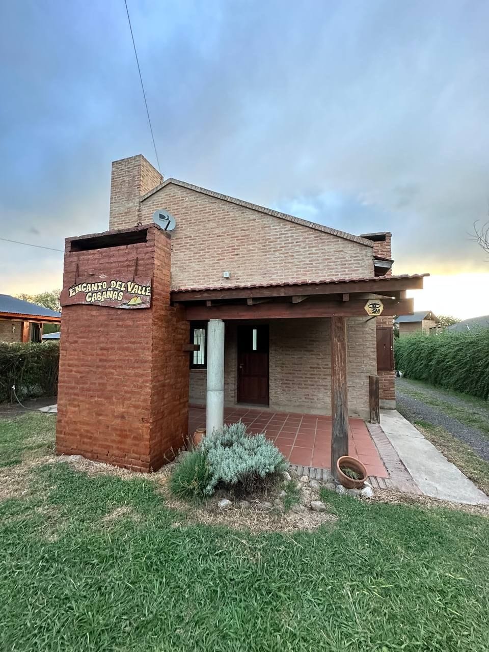 The cabaña displays a charming exterior with a combination of brick and stone. A covered porch with wooden beams provides a welcoming entryway, while grassy areas surround the building. The sky features soft, muted tones as evening approaches, adding a serene backdrop.