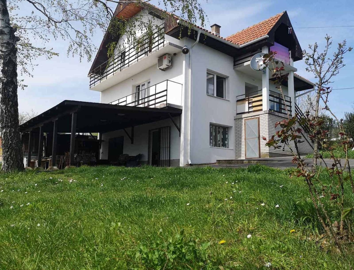 A modern two-story house with a white exterior is surrounded by a green grassy area. A covered patio area is visible on the ground level, offering a shaded outdoor space. Large windows on the upper floor provide natural light, enhancing the inviting atmosphere.