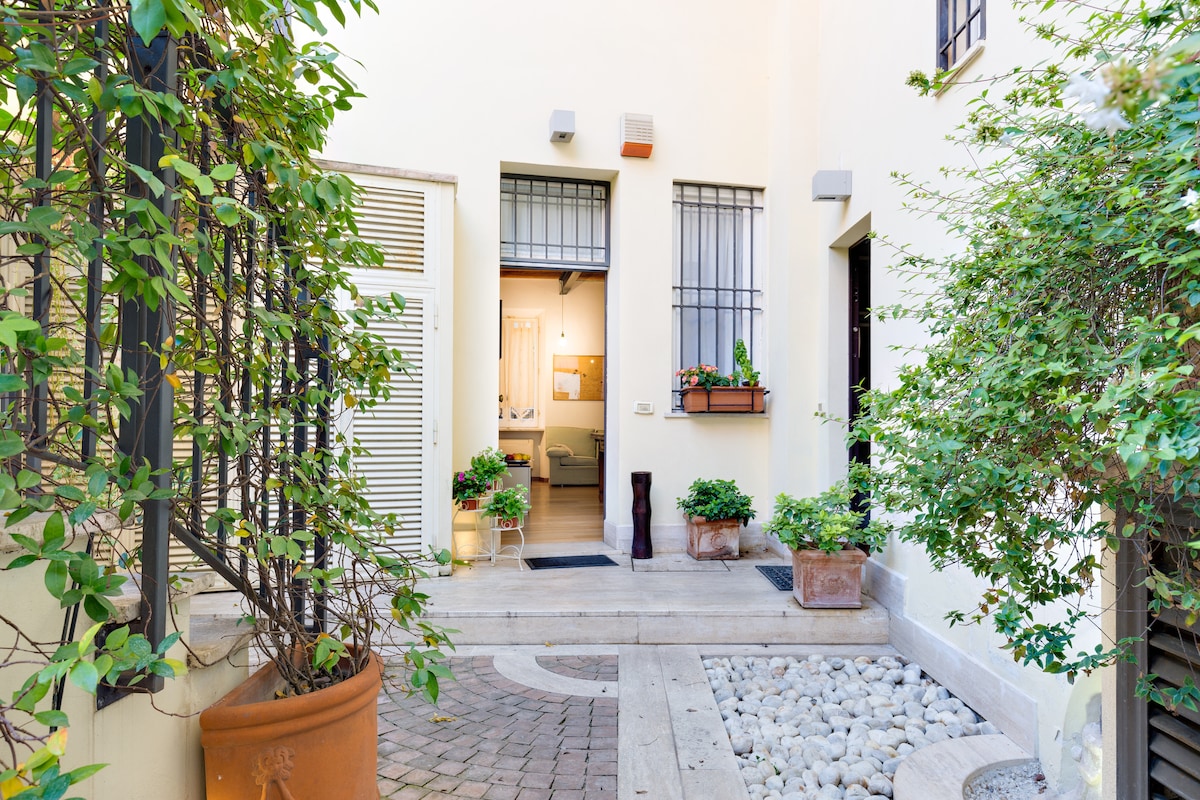 The courtyard is framed by lush greenery and flower pots, creating a welcoming entry. The stone pathway leads to two doors, hinting at the interior spaces. Light-colored walls offer a bright contrast, while decorative elements enhance the tranquil ambiance.