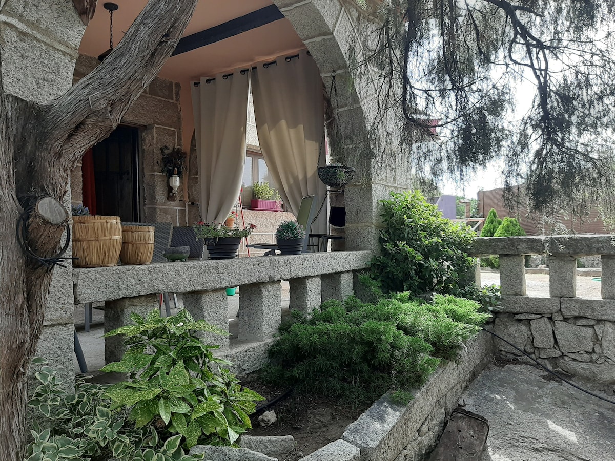 A stone cottage features a curved entrance with a stone railing leading to a shaded patio area. Potted plants are arranged on the railing, while light fabric curtains gently frame the entrance. Lush greenery surrounds the space, enhancing the garden's natural look.