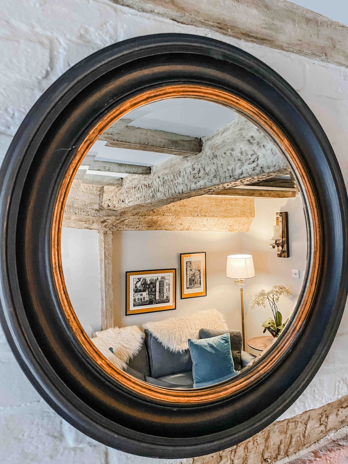 An oval mirror with a black and gold frame reflects the cozy sitting area. A two-seater sofa, adorned with cushions and a soft throw, is visible. The backdrop features original oak beams and framed artwork, while a lamp provides soft illumination, enhancing the inviting ambiance.