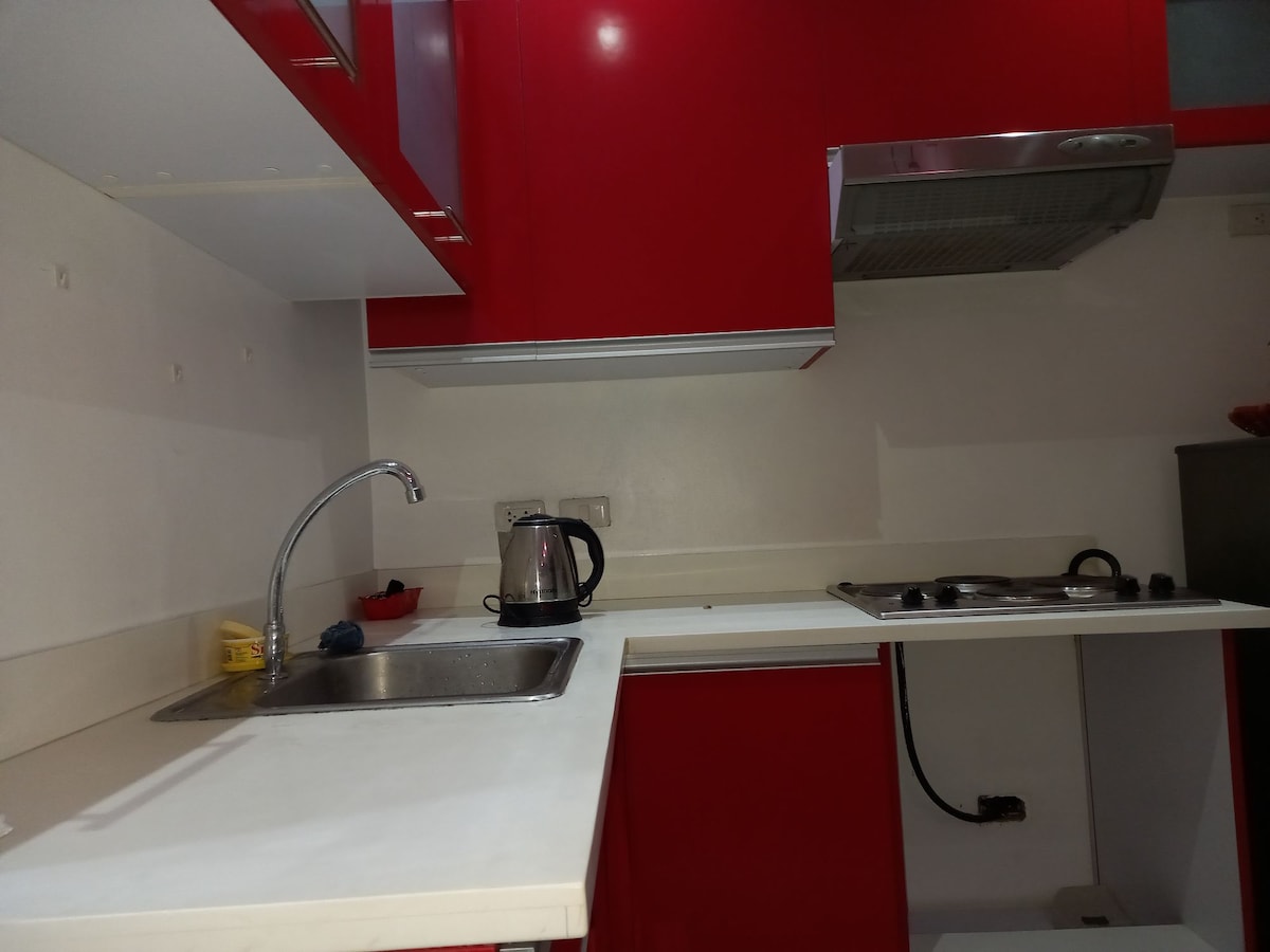 The kitchen features red cabinetry that adds a modern touch. A stainless-steel kettle sits on the countertop beside a sink. A stovetop is positioned adjacent to the sink, with a ventilation hood above, ensuring a functional and well-equipped cooking space.