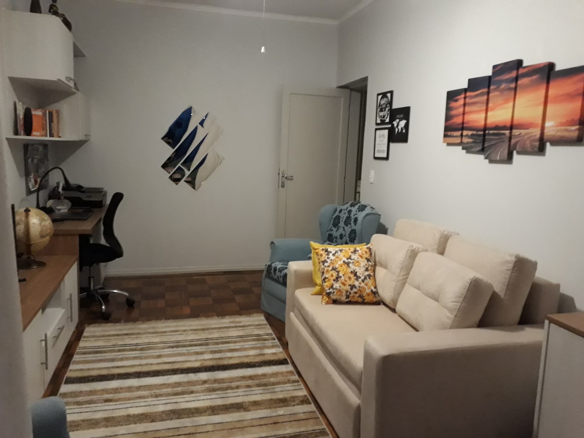A cozy living area is highlighted by a spacious sofa bed in a neutral tone, accompanied by two teal armchairs. A desk is positioned nearby, and wall art featuring vibrant landscapes is displayed. A striped rug adds texture to the wooden floor, enhancing the room's warmth.