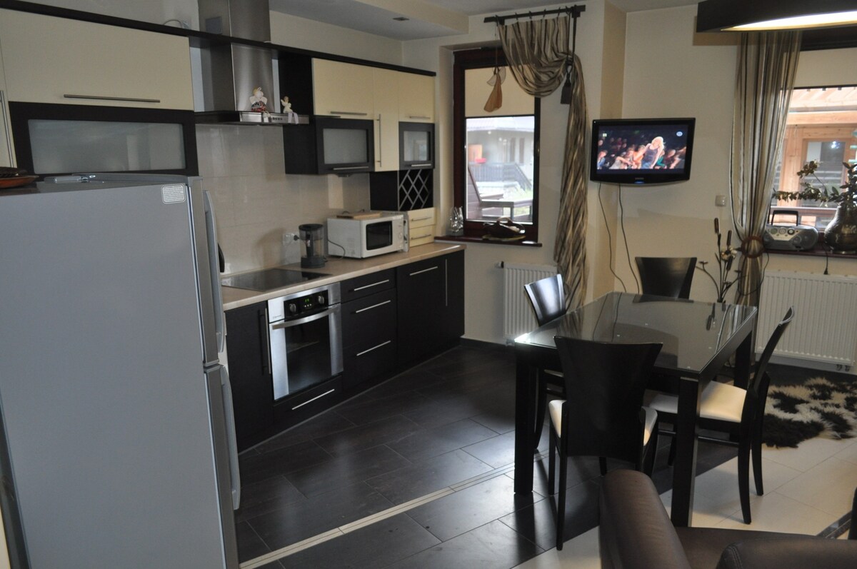 The contemporary kitchen is equipped with modern appliances, including a refrigerator, microwave, and oven. A dining table with black chairs is set in the center, while a television is mounted on the wall, enhancing the practicality of the space. Natural light enters through a window.