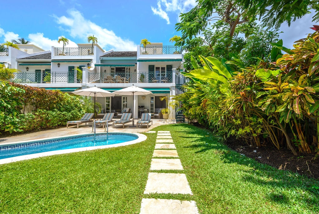 A spacious garden area is seen with a path leading to a private plunge pool. Sun loungers are arranged by the pool, accompanied by large umbrellas for shade. Lush greenery and colorful plants provide a vibrant backdrop to the townhouse's outdoor living space.