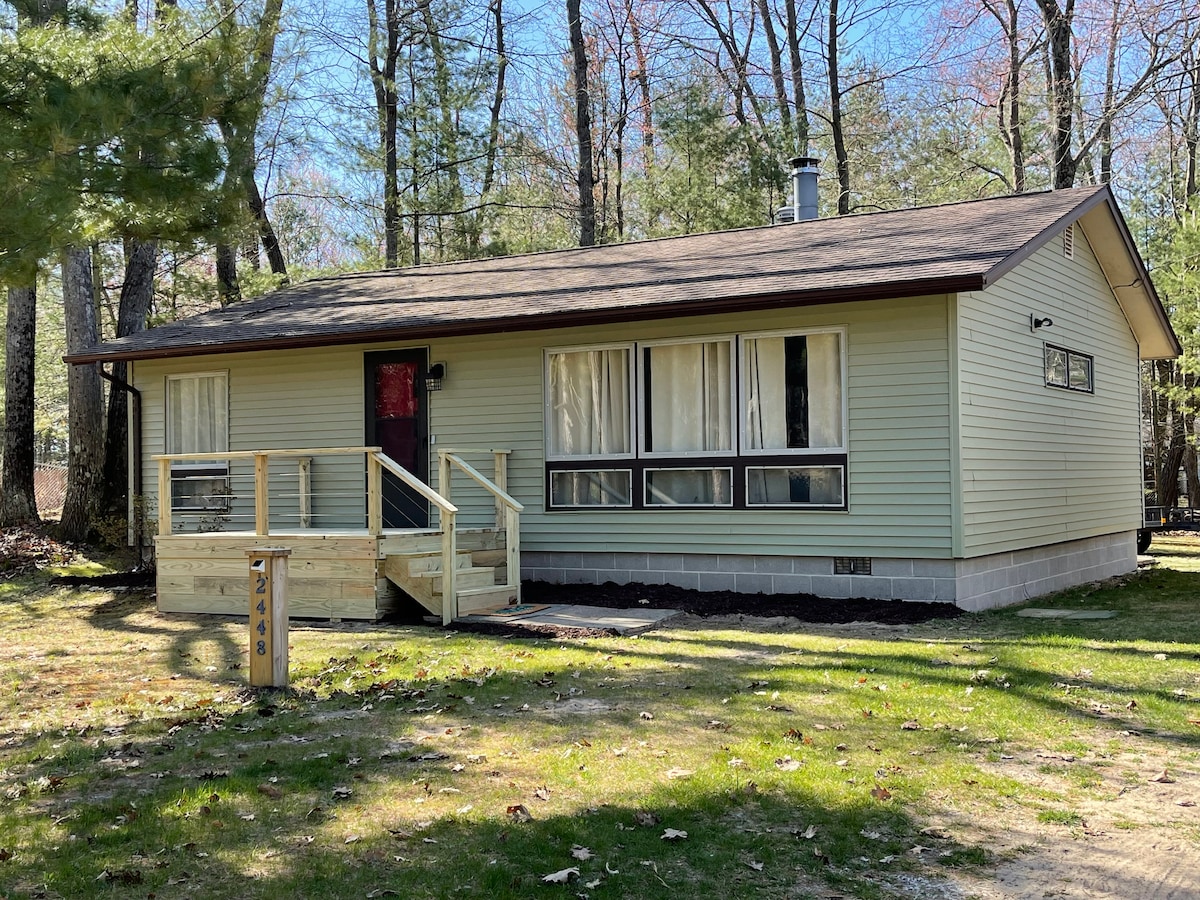 The front exterior of a single-story cottage is set within a wooded area. A wooden ramp leads to the entrance, with large windows allowing natural light into the living space. A calm green façade complements the surrounding trees, providing a serene environment.