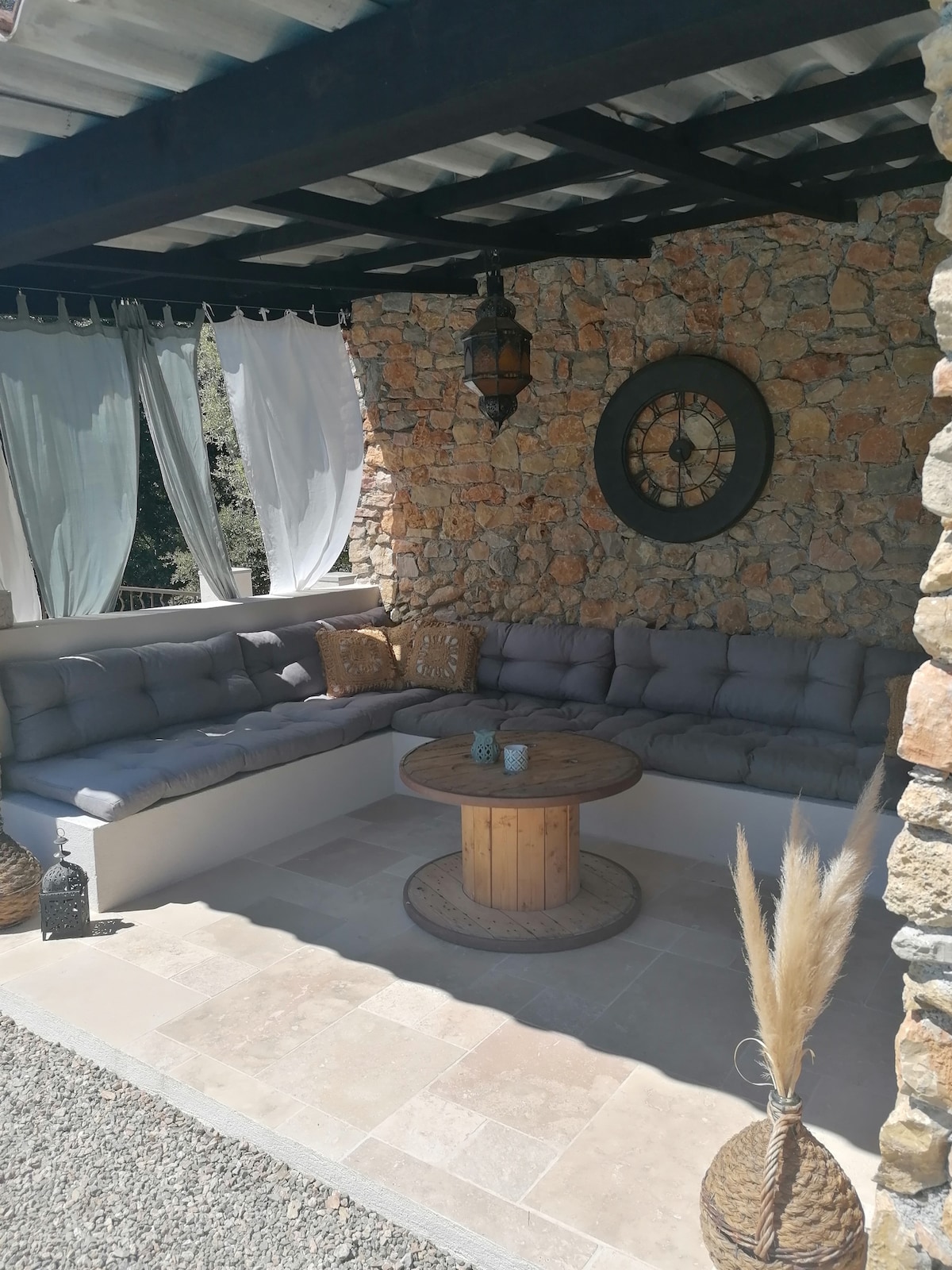 A shaded lounge area features a curved, cushioned seating arrangement with decorative throw pillows. A rustic wooden table sits in the center, and a stone wall adds texture to the setting. Soft drapes partially billow in the breeze, contributing to a relaxed ambiance.