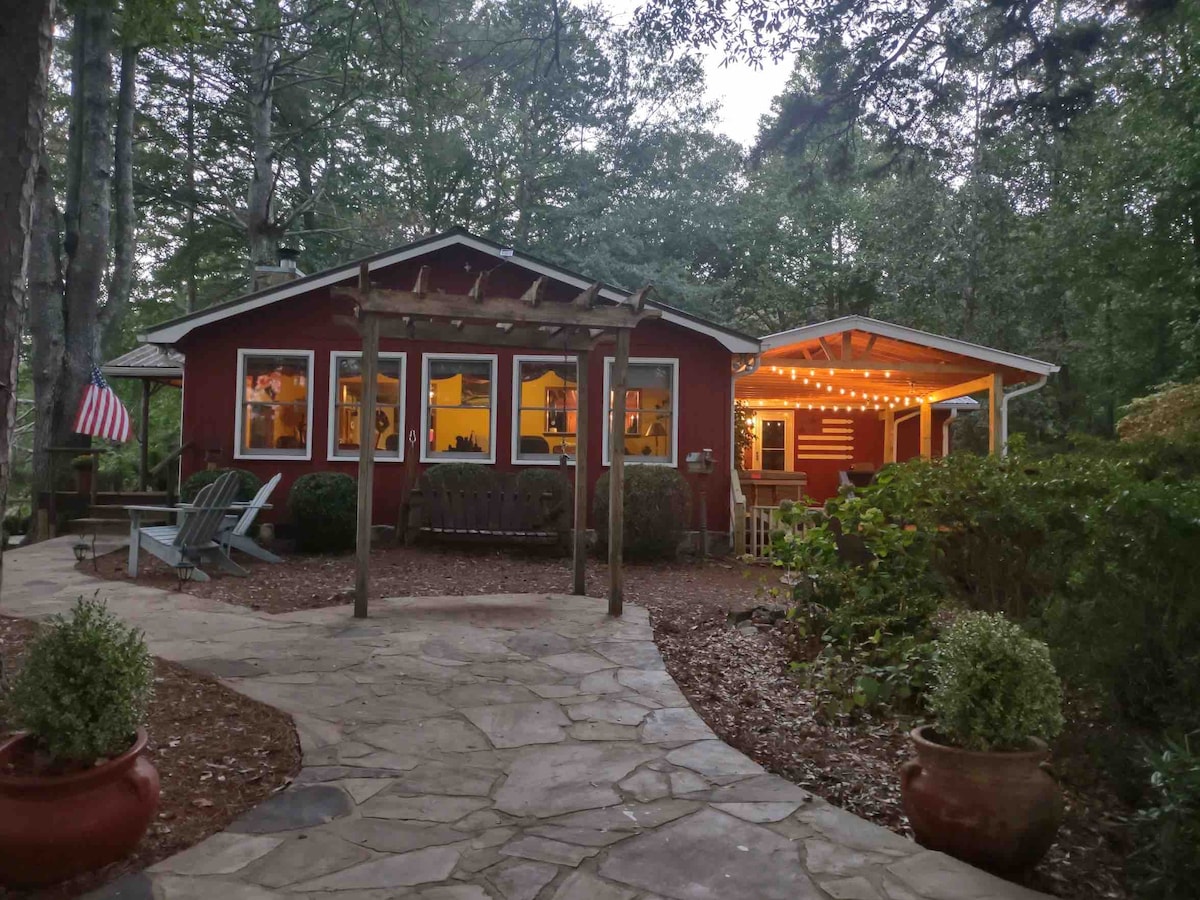 A charming single-family lake house features a rustic exterior with red siding, surrounded by lush greenery. A stone pathway leads to an inviting porch with warm string lights. Adirondack chairs provide outdoor seating amidst landscaped areas, enhancing the tranquil atmosphere.