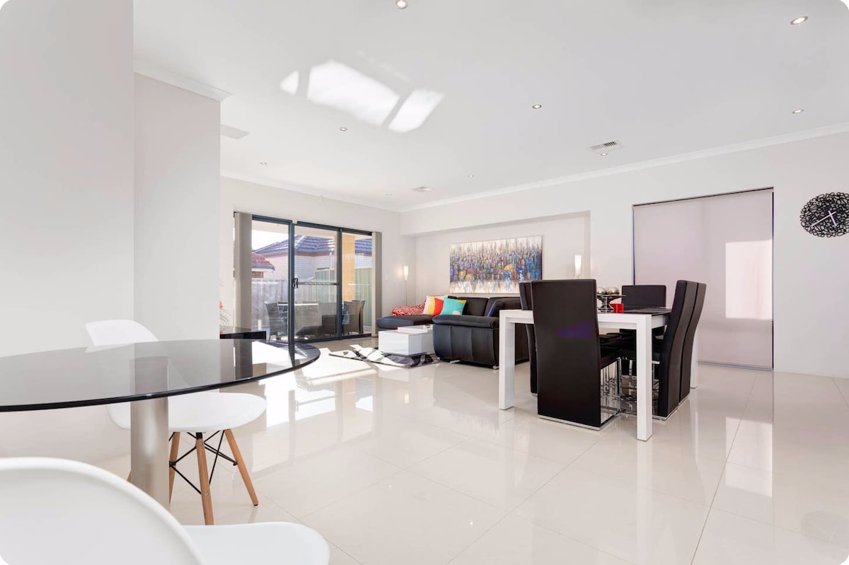An open-plan living and dining area features large glass doors allowing natural light to fill the space. Modern furnishings include a black leather sofa, a glass dining table with white chairs, and a colorful wall art piece that adds a touch of vibrancy.