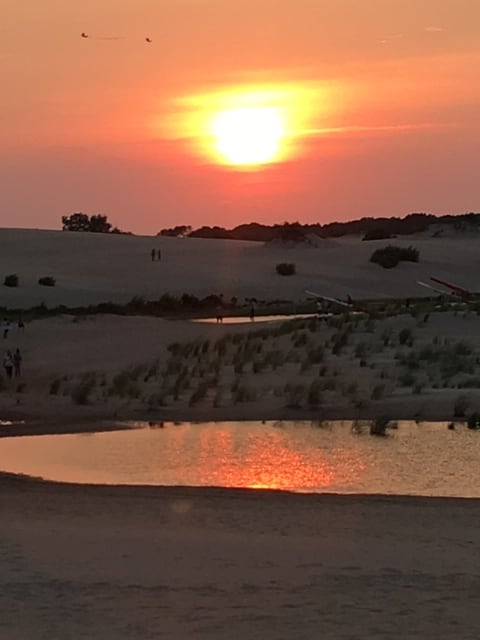 A tranquil sunset is captured over a sandy landscape, with vibrant orange and yellow hues reflecting on a calm body of water. People are visible in the distance, walking along the dunes, while sparse grass merges with the sand, enhancing the peaceful ambiance of the setting.