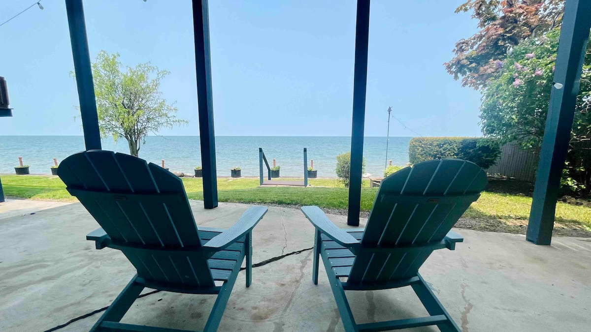 Two green Adirondack chairs are positioned on a covered patio, facing the serene Lake Ontario. The landscape features grassy areas and a clear view of the water, with a path leading down to the lake. Trees provide a gentle backdrop.
