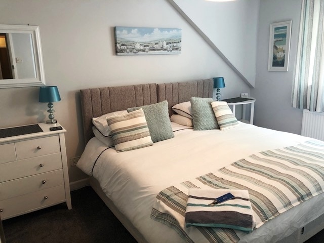 A spacious bedroom features a large bed with soft linens and decorative pillows. A light blue painting hangs above the headboard. Two bedside tables with lamps are positioned on either side, and a white dresser is situated in the corner.