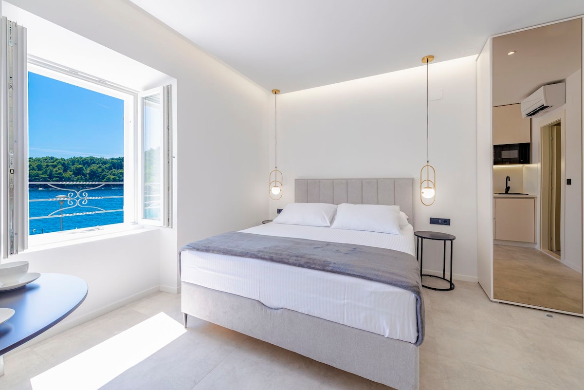 A bright and airy bedroom features a spacious bed with soft linens. A large window offers uninterrupted views of the sea, while two pendant lights hang elegantly beside the bed. A minimalist table is set against the wall, enhancing the room's clean design.