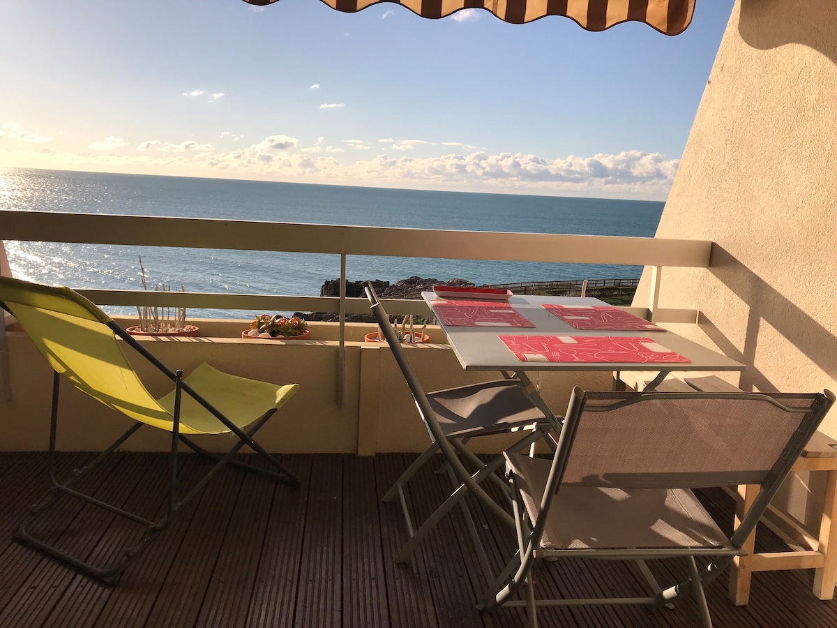 A balcony is visible, featuring a table with chairs and two reclining loungers. The ocean can be seen in the background, reflecting the sunlight. A canopy provides shade, and potted plants are arranged along the railing.