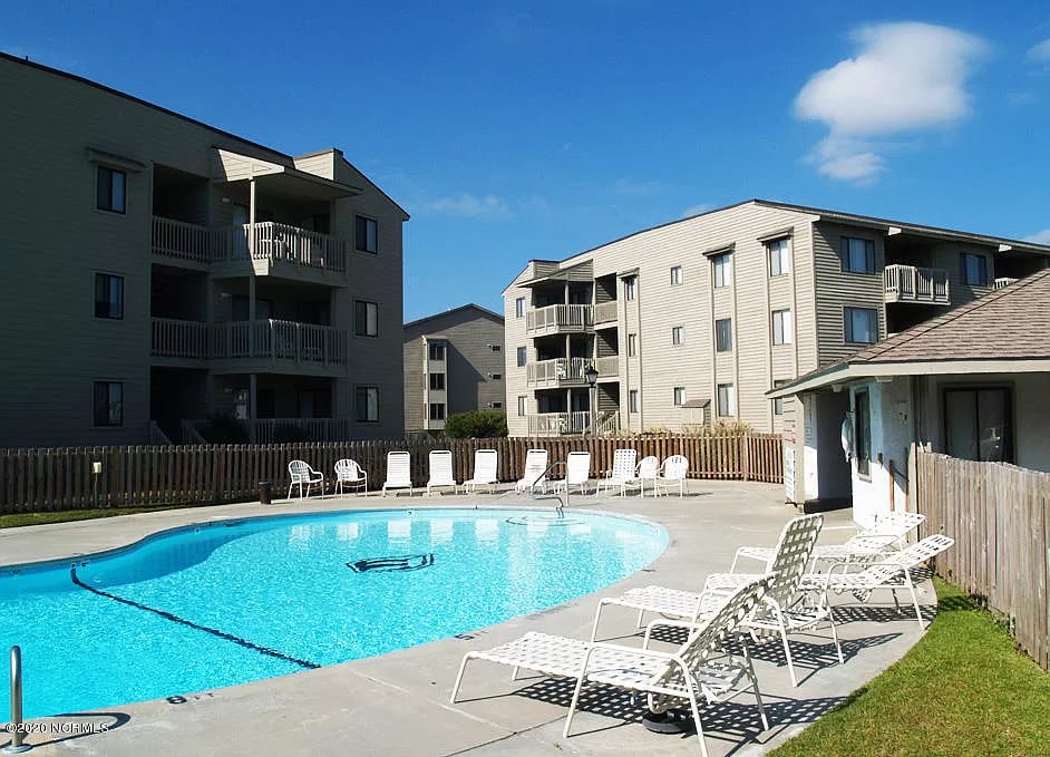 A bright outdoor pool area features a clear blue water surface surrounded by lounge chairs. The pool is located centrally among multistory buildings with balconies. A wooden fence provides an enclosed atmosphere, and a small building with a roof is visible at one side.