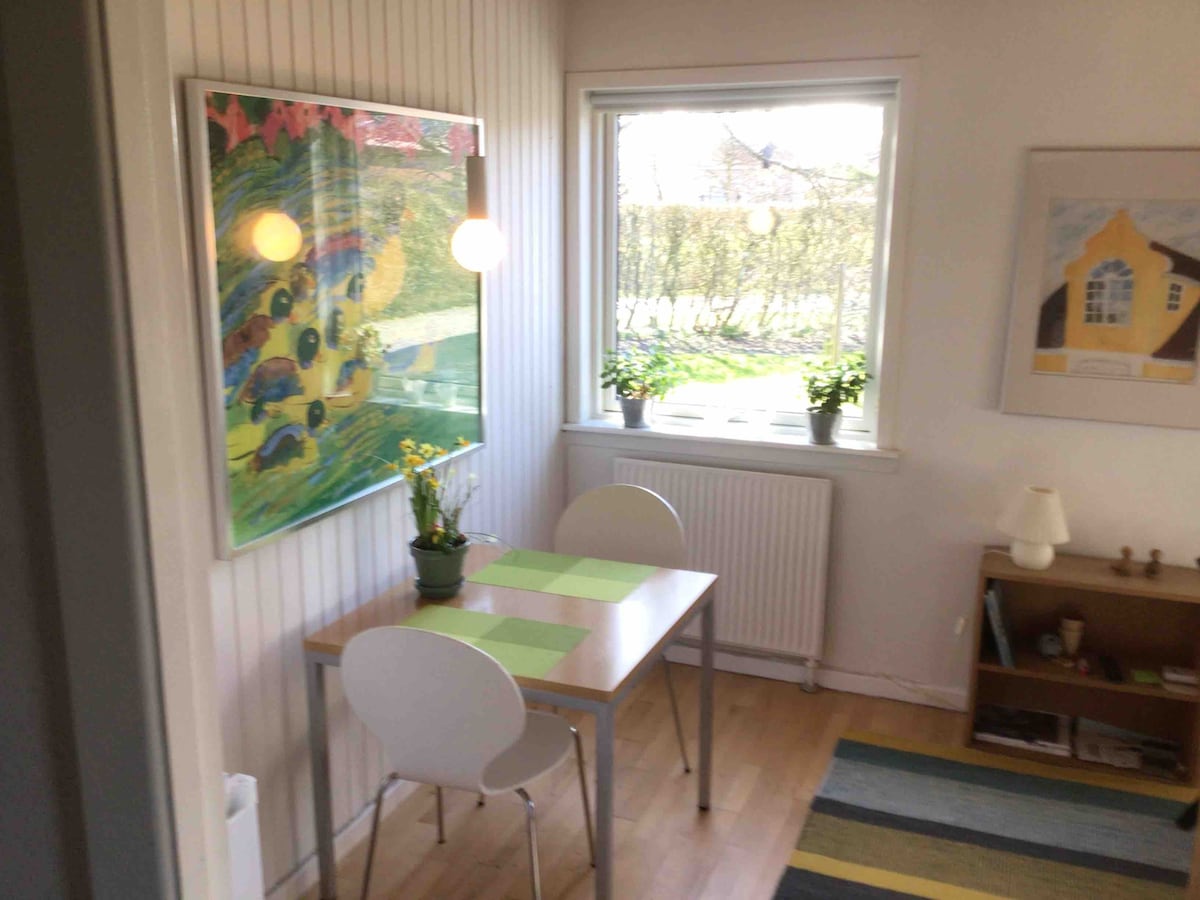 A small dining nook features a light wooden table accompanied by two white chairs. A colorful artwork adorns the wall, while potted plants sit on the windowsill, bringing a hint of nature indoors. Natural light streams through the window, illuminating the space.