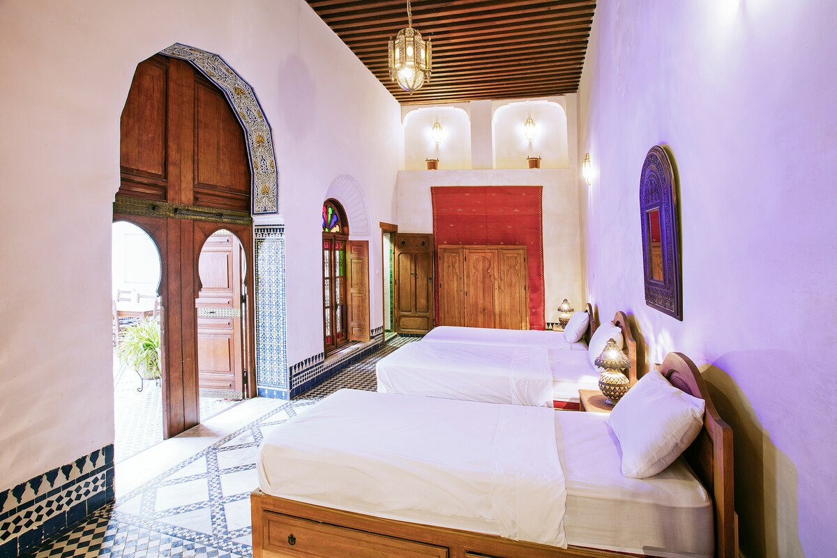 A spacious room with high ceilings features two standard single beds positioned against an elegant tiled floor. Large wooden doors open to a courtyard, while traditional lanterns provide ambient lighting. Decorative elements include a vibrant red wall and authentic Moroccan artistry enhancing the historical setting.