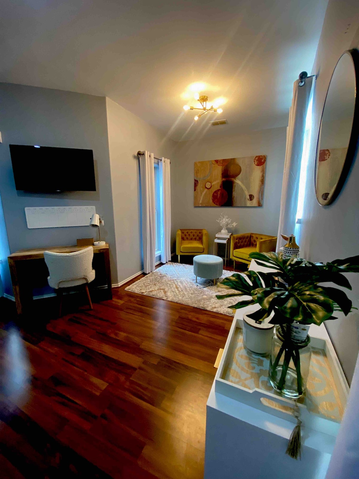 A welcoming living area features hardwood floors and soft blue-gray walls. A round rug anchors two accent chairs, complemented by a small table. A desk and television are positioned for convenience, while a potted plant adds a touch of greenery to the space.